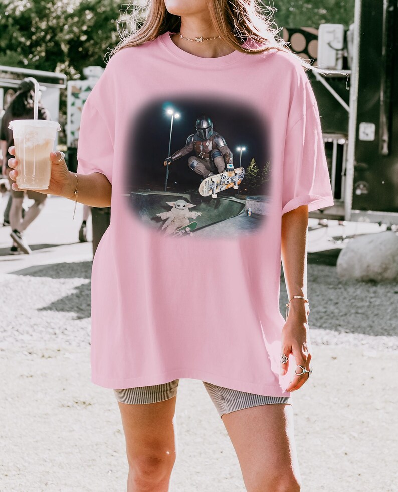 Op de afbeelding: Roze oversized T-shirt met een afbeelding van een personage met helm die in een skatepark skatet met een kleine groene alien. De afbeelding staat tegen een donkere achtergrond met heldere lichten.