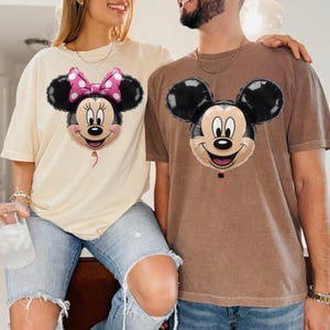 Peut inclure: Un t-shirt cr&egrave;me avec un motif de ballon Minnie Mouse portant un n&oelig;ud rose &agrave; pois et un t-shirt marron avec un motif de ballon Mickey Mouse. Les deux t-shirts pr&eacute;sentent des manches courtes et une encolure ronde classique.