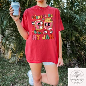 May include: Red t-shirt with the text "Counseling is My Jam" in a retro font. The graphic features a cartoon slice of bread and a jar of jam with arms and legs. The shirt is a comfortable, casual style.