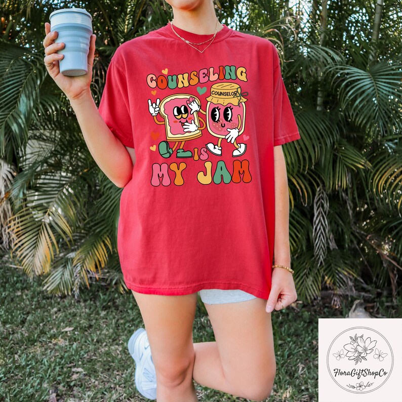 May include: Red t-shirt with the text "Counseling is My Jam" in a retro font. The graphic features a cartoon slice of bread and a jar of jam with arms and legs. The shirt is a comfortable, casual style.