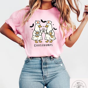 May include: Pink t-shirt featuring a cartoon design of two ducks dressed as nurses, holding medical supplies, with the word "GOOSEBUMPS" below. Black bat silhouettes are on the top. The shirt is made of a soft material.