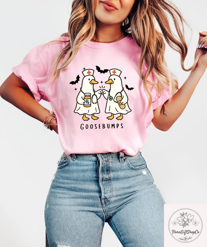 May include: Pink t-shirt featuring a cartoon design of two ducks dressed as nurses, holding medical supplies, with the word "GOOSEBUMPS" below. Black bat silhouettes are on the top. The shirt is made of a soft material.