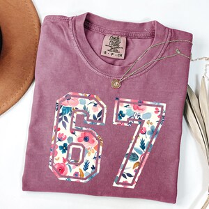 May include: A dusty rose t-shirt with the number "67" in a floral pattern. The shirt has a crew neck and a small tag near the collar. A gold necklace with a pendant is draped across the shirt. A brown hat is visible in the background.