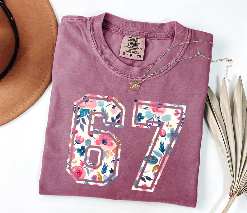 May include: A dusty rose t-shirt with the number "67" in a floral pattern. The shirt has a crew neck and a small tag near the collar. A gold necklace with a pendant is draped across the shirt. A brown hat is visible in the background.