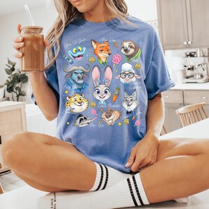 Puede incluir: Una camiseta de manga corta en color azul b&iacute;garo con un estampado gr&aacute;fico colorido de varios personajes animales animados de Zootopia, incluidos Judy Hopps, Nick Wilde y Flash, dispuestos en un collage con peque&ntilde;os iconos como zanahorias.