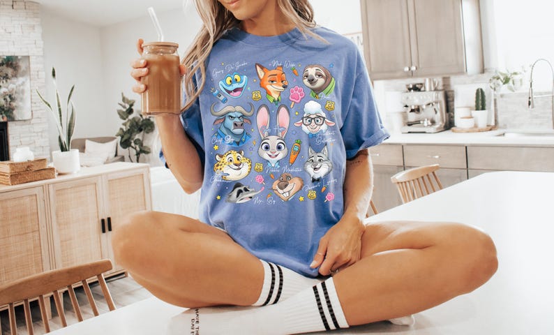 Puede incluir: Una camiseta de manga corta en color azul b&iacute;garo con un estampado gr&aacute;fico colorido de varios personajes animales animados de Zootopia, incluidos Judy Hopps, Nick Wilde y Flash, dispuestos en un collage con peque&ntilde;os iconos como zanahorias.
