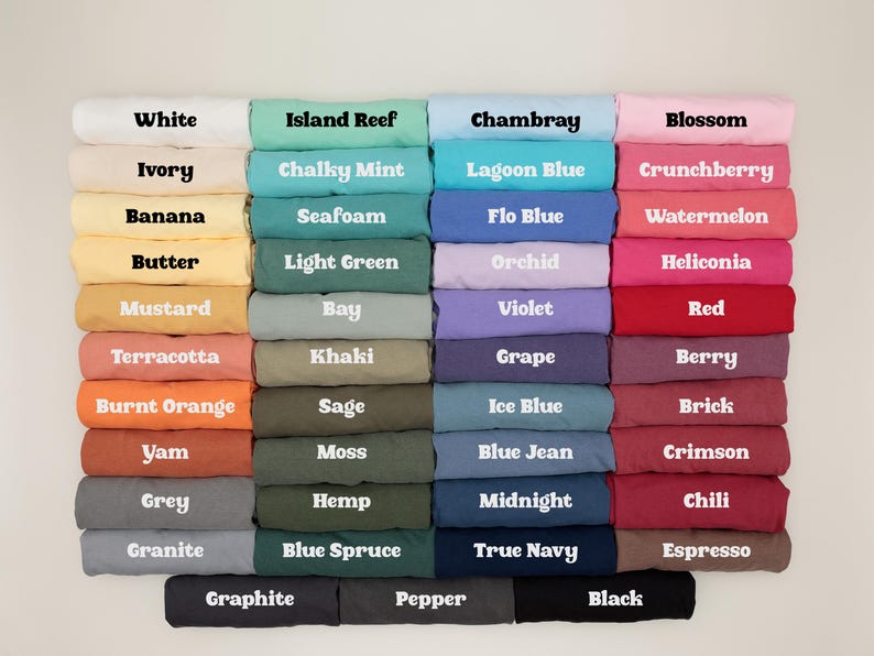 May include: A display of folded t-shirts in various colors, including white, blue, pink, and green. Each shirt is labeled with its color name in white text, such as "White," "Chambray," and "Blossom." The shirts are neatly stacked.