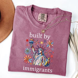 May include: A mauve short-sleeve t-shirt featuring a graphic of the Statue of Liberty surrounded by colorful flowers, stars, and fireworks. The text "built by immigrants" is printed in white above and below the graphic. The shirt has a crew neck.