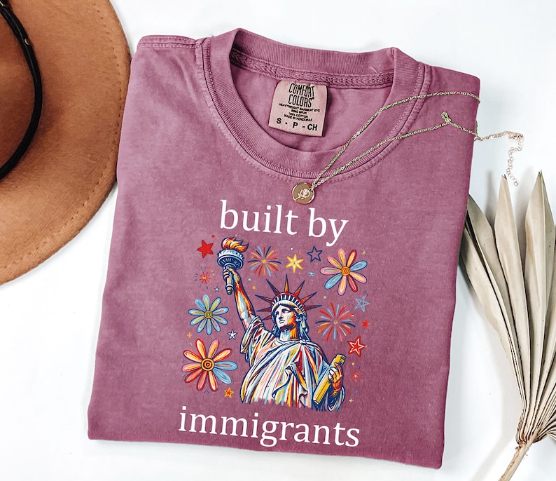 May include: A mauve short-sleeve t-shirt featuring a graphic of the Statue of Liberty surrounded by colorful flowers, stars, and fireworks. The text "built by immigrants" is printed in white above and below the graphic. The shirt has a crew neck.