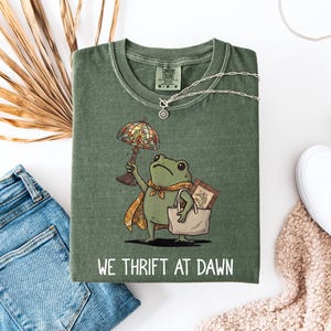 Puede incluir: Una camiseta verde oliva con el gráfico de una rana que lleva una capa estampada, sostiene una lámpara de vidrieras y lleva un bolso con un cuadro enmarcado dentro. El texto WE THRIFT AT DAWN está impreso en blanco bajo la ilustración.