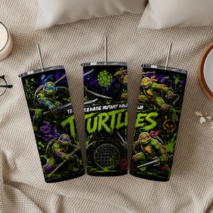 May include: Three cylindrical tumblers with metal straws featuring Teenage Mutant Ninja Turtles graphics. The design includes green turtles with colored masks, weapons, and the text "TEENAGE MUTANT NINJA TURTLES" in bright green against a dark background.