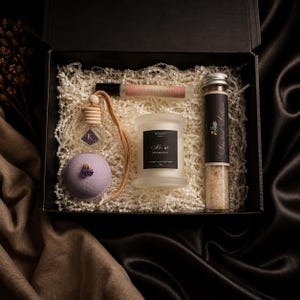 May include: A black gift box filled with white crinkle paper containing a glass roller bottle labelled Golden Hour, a small hanging car diffuser, a lavender bath bomb with dried flowers, a white candle labelled No. 67, and a glass tube of bath salts.