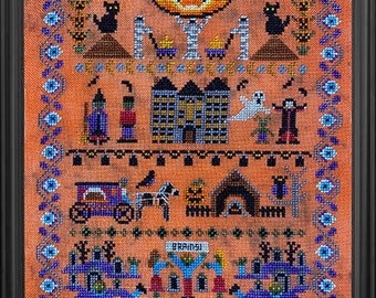 May include: A cross-stitch pattern featuring a grinning orange jack-o'-lantern with a black cat, a haunted house, and other Halloween imagery. The pattern is framed in a black frame.