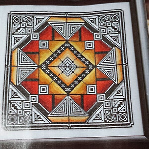 Peut inclure: Une œuvre d'art au point de croix encadrée présentant un motif géométrique avec un losange central. Le motif comprend des carrés, des triangles et des motifs complexes dans des tons de rouge, orange, jaune, noir et blanc. L'œuvre est encadrée dans un cadre marron foncé.