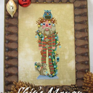 May include: A cross-stitch pattern of a woodland nutcracker with a blue owl on its head, a squirrel at its feet, and a lantern hanging from its arm. The nutcracker is wearing a red and green plaid shirt and has a white beard. The background is a light beige fabric. The image is framed in a brown wooden frame with a gold trim. The text "Woodland Nutcracker" is at the top of the frame and "Ship's Manor" is at the bottom.