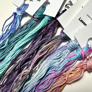 May include: A close-up of several skeins of embroidery floss in various shades of blue, purple, pink, and peach. The floss is arranged in a fan shape, with the ends of the skeins gathered together at the top. The skeins are labeled with the brand name "DMC" and the color numbers.