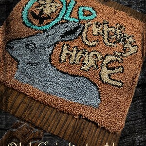 May include: A brown and tan needlepoint wall hanging with the words "Old Cricketty Hare" in a whimsical font. The design features a stylized hare with a circle around its head.
