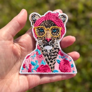 May include: An embroidered patch featuring a leopard wearing a bright pink headwrap, yellow-rimmed glasses, and a pink shirt with a floral pattern of red roses and blue leaves. The leopard has blue eyes and a white muzzle, with a white border edge.