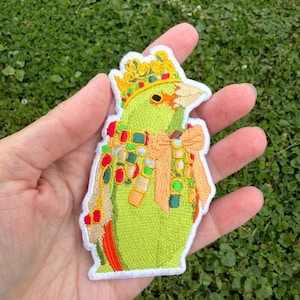 May include: An embroidered patch featuring a bright green parrot wearing a yellow crown with red and blue accents. The parrot has a light orange beak, a tan scarf with colorful square patterns, and a large tan bow tied around its neck.