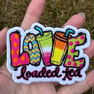 Puede incluir: Un parche bordado con la palabra LOVE donde las letras O y V están reemplazadas por dos vasos de bebida coloridos con pajitas. La letra L es rosa con puntos naranjas, la letra E es verde con puntos rosas, y el texto loaded tea está debajo.