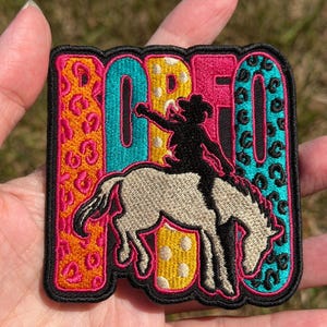 May include: A rectangular embroidered patch featuring the word RODEO in bold letters. The letters are filled with orange, teal, and yellow animal print patterns. A black silhouette of a rider on a bucking horse is centered over the text.