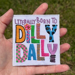 Könnte beinhalten: Quadratischer, gestickter Aufnäher mit dem Text "LITERALLY BORN TO DILLY DALLY". Die Buchstaben sind in verschiedenen Farben gehalten, darunter Orange, Pink, Blau, Grün und Lila. Der Aufnäher hat einen weißen Rand und dekorative Blumen- und Musterdetails.