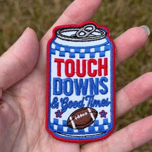 Puede incluir: Un parche bordado con forma de lata de bebida con un patrón de cuadros rojo, blanco y azul. Presenta el texto en rojo y azul "TOUCH DOWNS & Good Times" sobre un gráfico de un balón de fútbol americano marrón y dos estrellas azules.