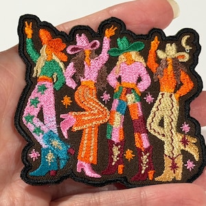 May include: An embroidered patch featuring four figures wearing cowboy hats and boots. The figures are dressed in colorful, patterned clothing including pink, orange, green, and yellow hues. Small star and moon icons are scattered around.