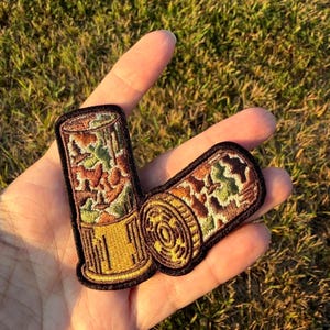 May include: An embroidered patch featuring two shotgun shells with a camouflage pattern in shades of green, brown, and tan. The base of the shells is a metallic gold color with vertical lines, and the entire patch is outlined in black thread.