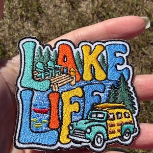 May include: An embroidered patch featuring the text LAKE LIFE in colourful block letters. The design includes a wooden dock with two chairs, a red canoe on blue water, green pine trees, and a teal and yellow station wagon with a surfboard.