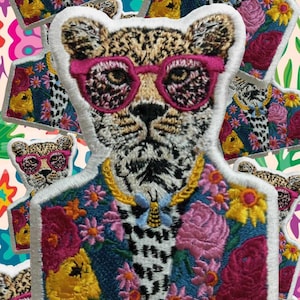 Puede incluir: Parche bordado con un leopardo que lleva gafas de sol rosas y una chaqueta floral. El leopardo tiene un collar de cadena dorada y un colgante de abeja. La chaqueta está decorada con flores rosas, amarillas y rojas. El parche tiene un borde blanco.