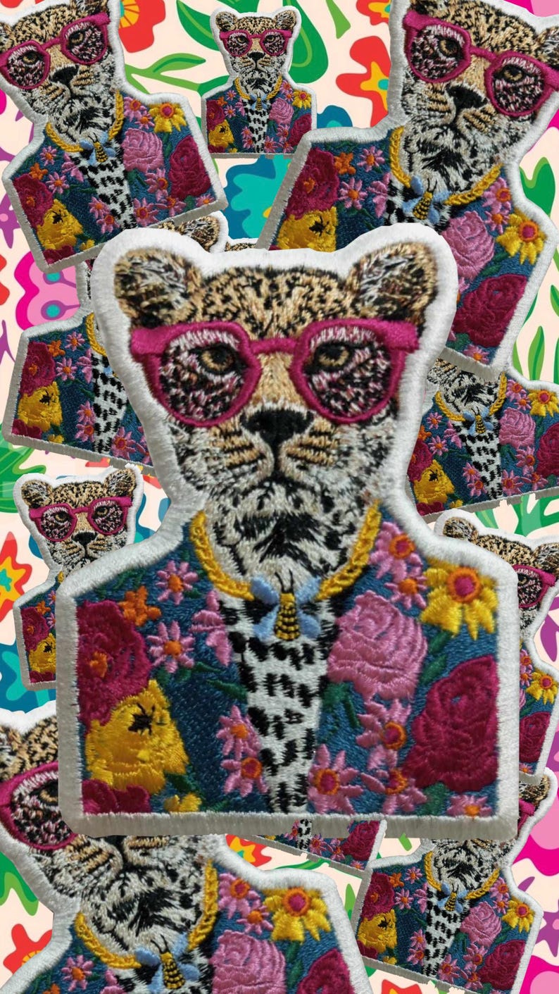 May include: Embroidered patch of a leopard wearing pink sunglasses and a floral jacket. The leopard has a gold chain necklace and a bee pendant. The jacket is decorated with pink, yellow, and red flowers. The patch has a white border.