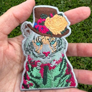 May include: An embroidered patch featuring a white tiger with bright blue eyes and a pink nose. The tiger wears a brown top hat adorned with a large yellow flower and smaller pink blossoms. The tiger is depicted wearing a green shirt with a dark tie.