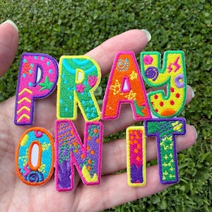 Pode incluir: Um emblema bordado com as palavras PRAY ON IT em letras vibrantes e multicoloridas. Cada letra apresenta um padrão único, incluindo pontos, espirais, estrelas e ziguezagues em tons de rosa, amarelo, verde, azul, laranja e roxo.