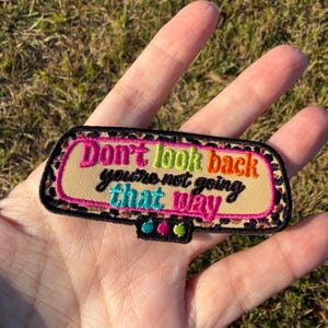 Puede incluir: Un parche bordado rectangular con forma de espejo retrovisor y borde de estampado de leopardo. El centro beige presenta el texto "Don't look back you're not going that way" en hilos de colores rosa, verde, naranja, negro y turquesa.