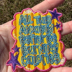 May include: An embroidered patch with a yellow background and a pink border. The text reads "ALL THIS ANXIETY BETTER BE BURNING CALORIES" in blue letters. The design includes three purple stars and small white accents around the edges.