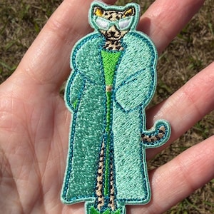 May include: An embroidered patch featuring a leopard character wearing white sunglasses, a bright green top, and a long, light teal coat. The character has a spotted tail and is wearing green shoes. The patch has a thick teal border.