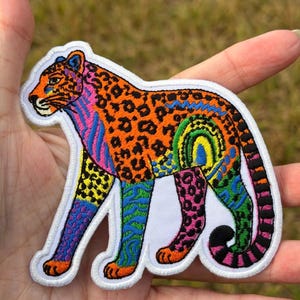 May include: An embroidered patch in the shape of a leopard featuring a vibrant, multi-colored pattern. The design includes orange, blue, pink, green, and yellow sections with leopard spots, stripes, and a rainbow arc on the animal's flank.