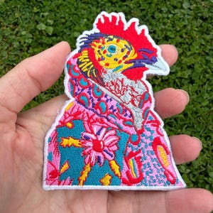 May include: An embroidered patch featuring a stylized rooster head with a bright red comb, a yellow and blue patterned face, and a white beak. The neck and body area display a vibrant, multi-colored abstract design with pink, blue, and yellow floral and geometric shapes.