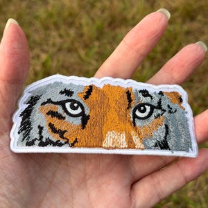 Könnte beinhalten: Ein bestickter Aufnäher mit den detaillierten Augen und dem Fell eines Tigers. Das Design zeigt leuchtend orangefarbene, graue und schwarze Fäden mit einem weißen Rand und fängt den intensiven Blick des Tieres in einer rechteckigen Form ein.