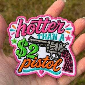 Peut inclure: Un écusson brodé avec un fond blanc et une bordure rose. Il présente le texte 'hotter THAN A $2 pistol' en rose, bleu sarcelle et orange. Un revolver gris avec une poignée à motif léopard rose et noir est centré sur l'écusson.