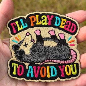 May include: An embroidered patch featuring a grey and cream-coloured opossum lying on its back with its tongue out and an X over one eye. The text "I'LL PLAY DEAD TO AVOID YOU" is stitched in colourful letters above and below the opossum.