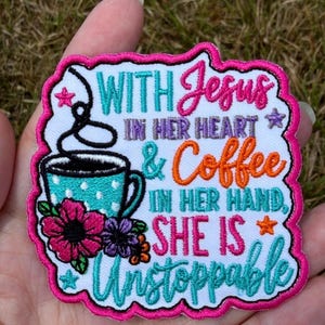 Puede incluir: Un parche bordado blanco con borde rosa intenso que presenta una taza de café turquesa con lunares, flores coloridas y el texto: WITH Jesus IN HER HEART & Coffee IN HER HAND, SHE IS Unstoppable. El diseño incluye pequeños detalles de estrellas.