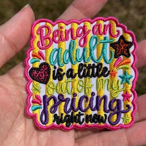 May include: A square embroidered patch with a scalloped pink border featuring yellow and white vertical stripes. The text reads Being an adult is a little out of my pricing right now in pink, teal, black, and purple thread with heart and star accents.