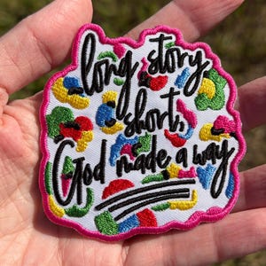 May include: An embroidered patch with a white background and a bright pink border. The patch features colorful abstract shapes in yellow, green, blue, and red, with black embroidered text that reads: long story short God made a way.