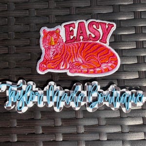 May include: A pink and orange embroidered patch featuring a tiger with the word "EASY" above it. Below is a checkered design with the words "Taylor Made Boutique" in blue script.