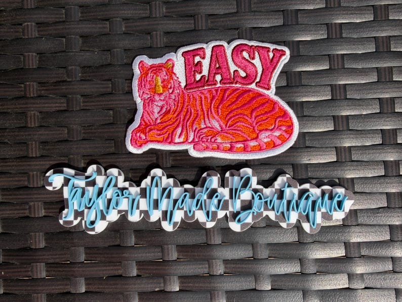 May include: A pink and orange embroidered patch featuring a tiger with the word "EASY" above it. Below is a checkered design with the words "Taylor Made Boutique" in blue script.