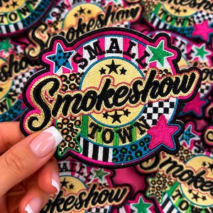 May include: A circular embroidered patch featuring the text Small Smokeshow Town in black and gold lettering. The design includes a yellow center, black and white checkered patterns, leopard print accents, and bright pink, blue, and green stars.