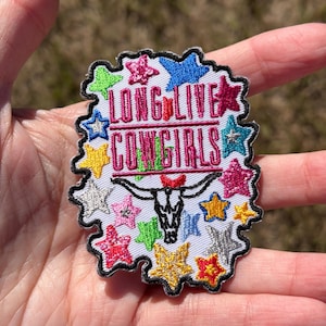May include: An embroidered patch with a white background featuring the text LONG LIVE COWGIRLS in pink, a black steer skull graphic, and numerous multi-colored stars in shades of blue, yellow, green, pink, and silver with a black border.