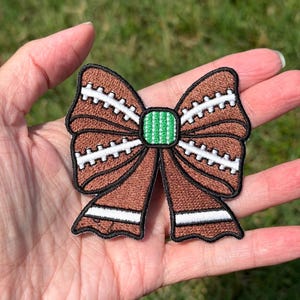 May include: A brown embroidered patch shaped like a bow, featuring white stitching that mimics the laces of an American football. The center of the bow has a small green square with white vertical lines, and the tails have white horizontal stripes.